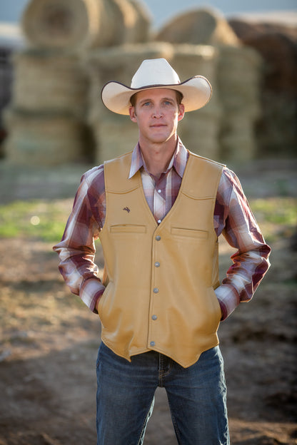 Man wearing a cowboy hat, yellow vest, and plaid shirt standing in front of hay bales.